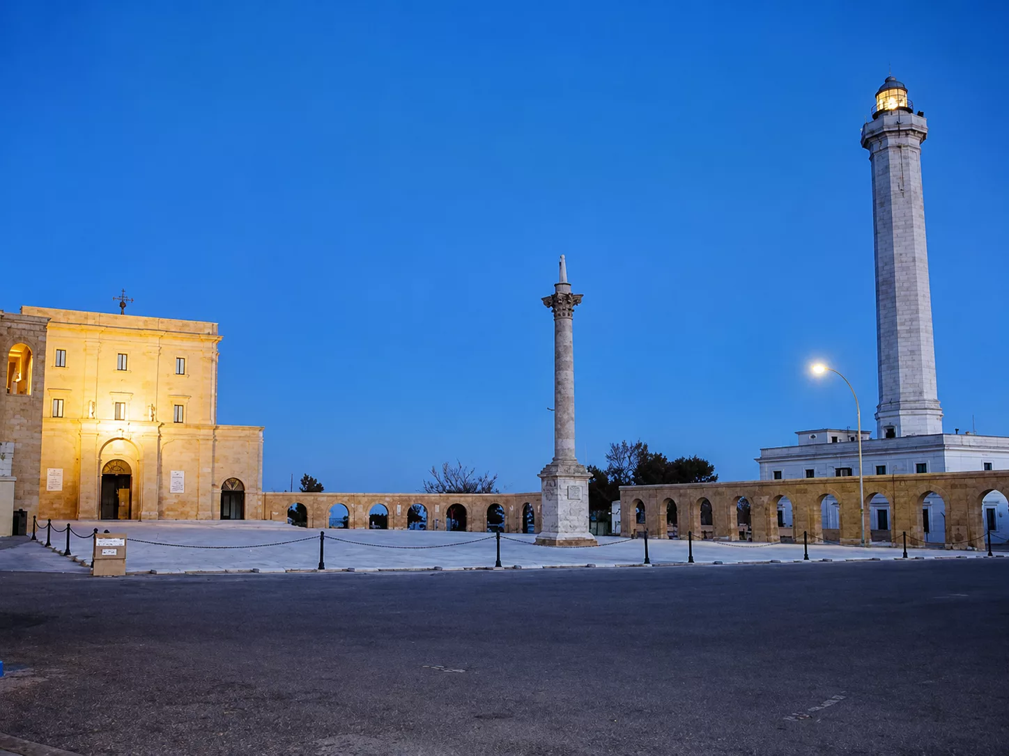 Veduta serale del Santuario di Santa Maria di Leuca, con la colonna monumentale e il faro illuminato.